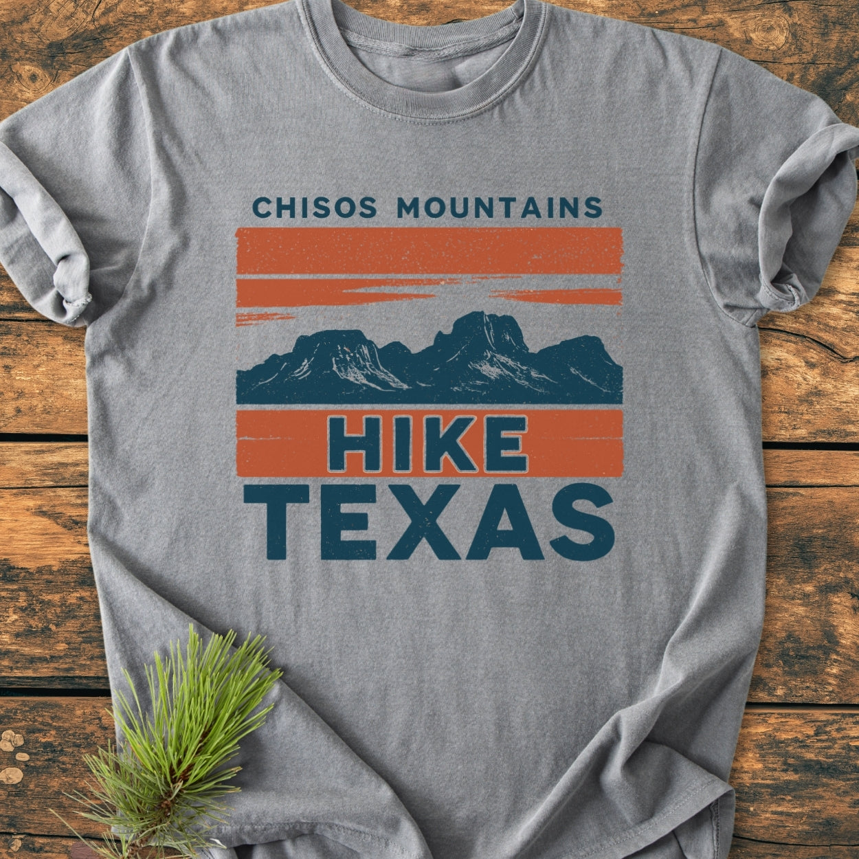 Chisos Mountains Texas - Vintage Wash Tee on wooden surface, showcasing an orange and dark blue mountain design with “HIKE TEXAS.” A pine sprig accents this soft, relaxed-fit, heather-gray shirt.