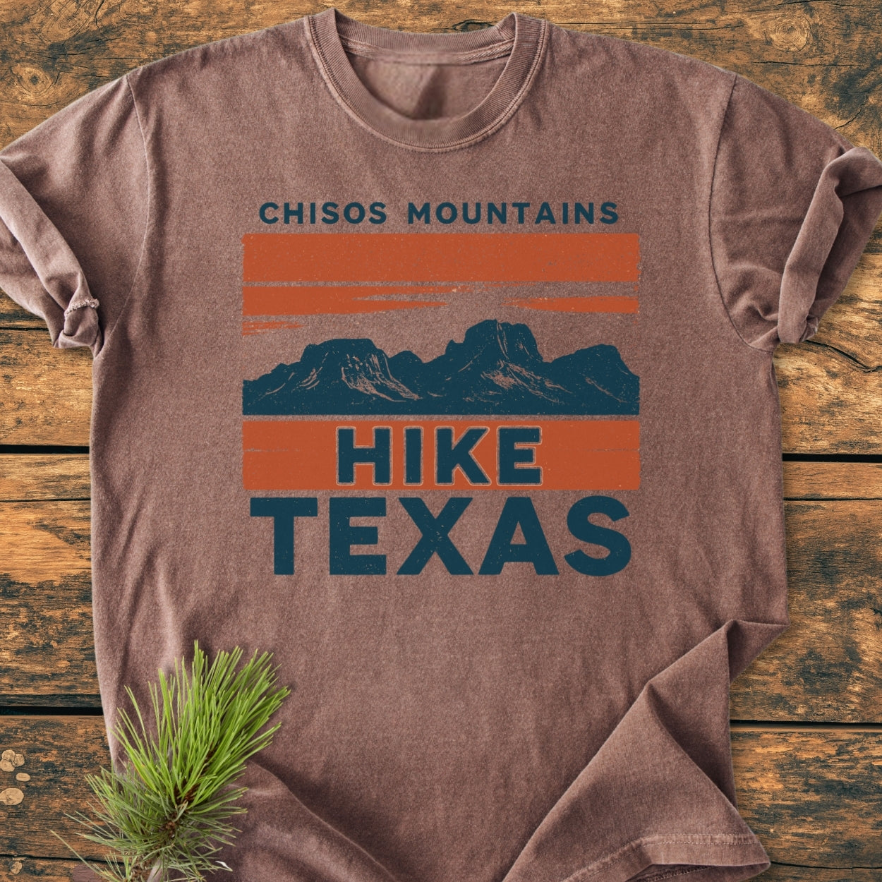 Chisos Mountains Texas - Vintage Wash Tee features a heathered-brown design with a mountain graphic. Laid on wood, it highlights the shirt's rolled sleeve and pine sprig accent, embodying rugged style.