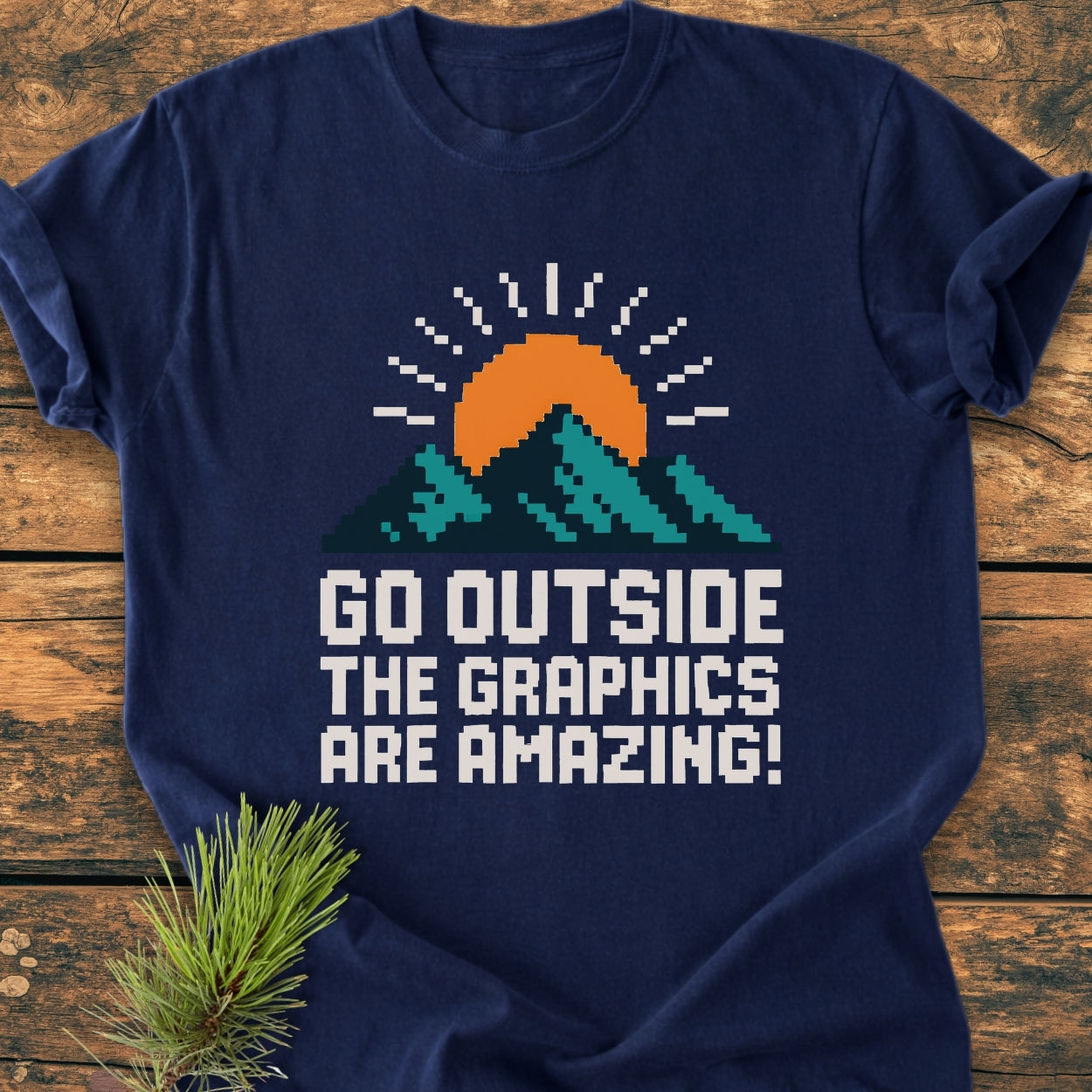 Go Outside Pixelart Mountain - Vintage Wash Tee features an 8-bit teal mountains design with an orange sun, laid on a wooden surface, highlighting its retro, cozy style and relaxed fit.