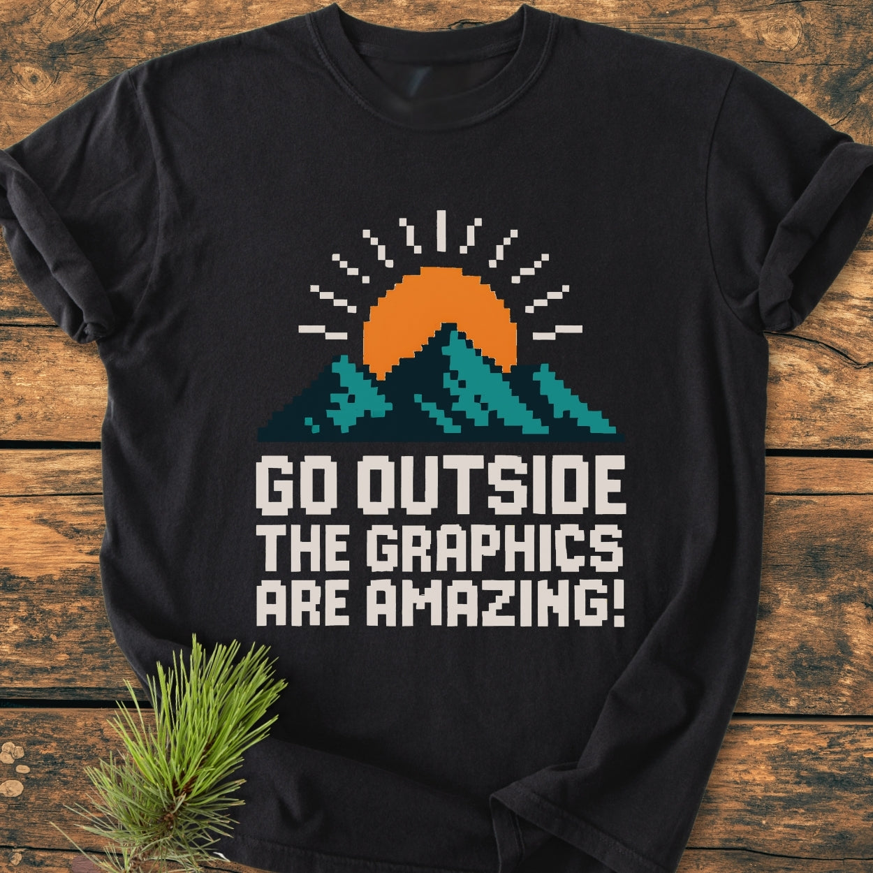 Go Outside Pixelart Mountain - Vintage Wash Tee features a retro pixel-art graphic of teal mountains and an orange sun, printed on a black tee, laid on a wood-plank background with pine sprig accents.