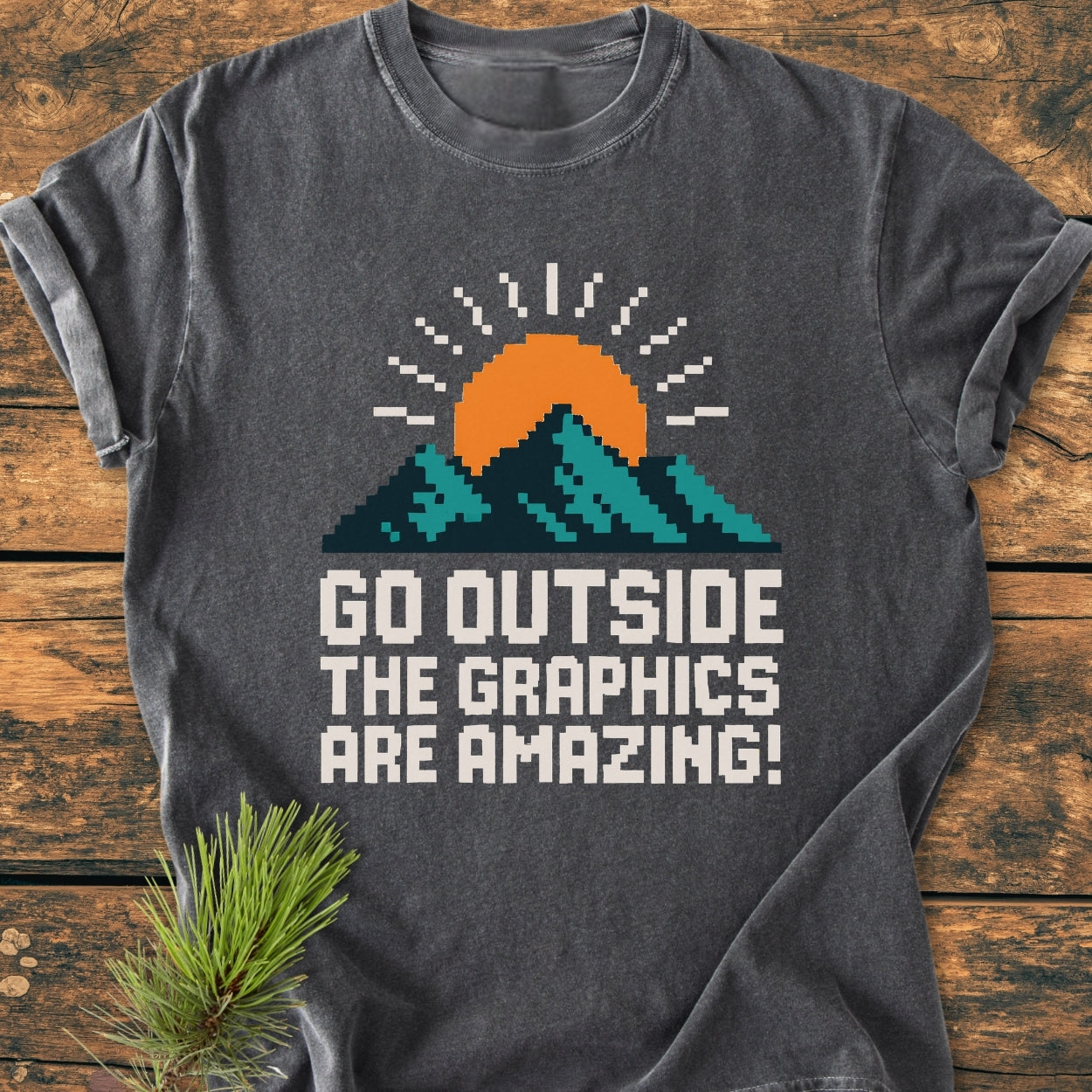 Go Outside Pixelart Mountain - Vintage Wash Tee featuring an 8-bit mountain and sun graphic on a dark gray background, laid on a wooden plank with a pine sprig nearby.