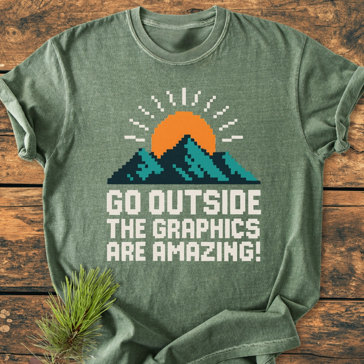 Heather-green Go Outside Pixelart Mountain - Vintage Wash Tee featuring an 8-bit sunrise with mountains and a pixelated slogan, laid flat on a wooden surface.
