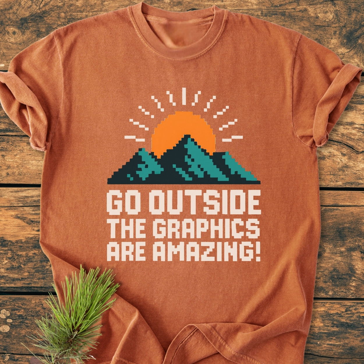 Rust-orange Go Outside Pixelart Mountain - Vintage Wash Tee featuring an 8-bit sunrise and mountain design with a slogan, laid on a wooden backdrop with a pine sprig accent.