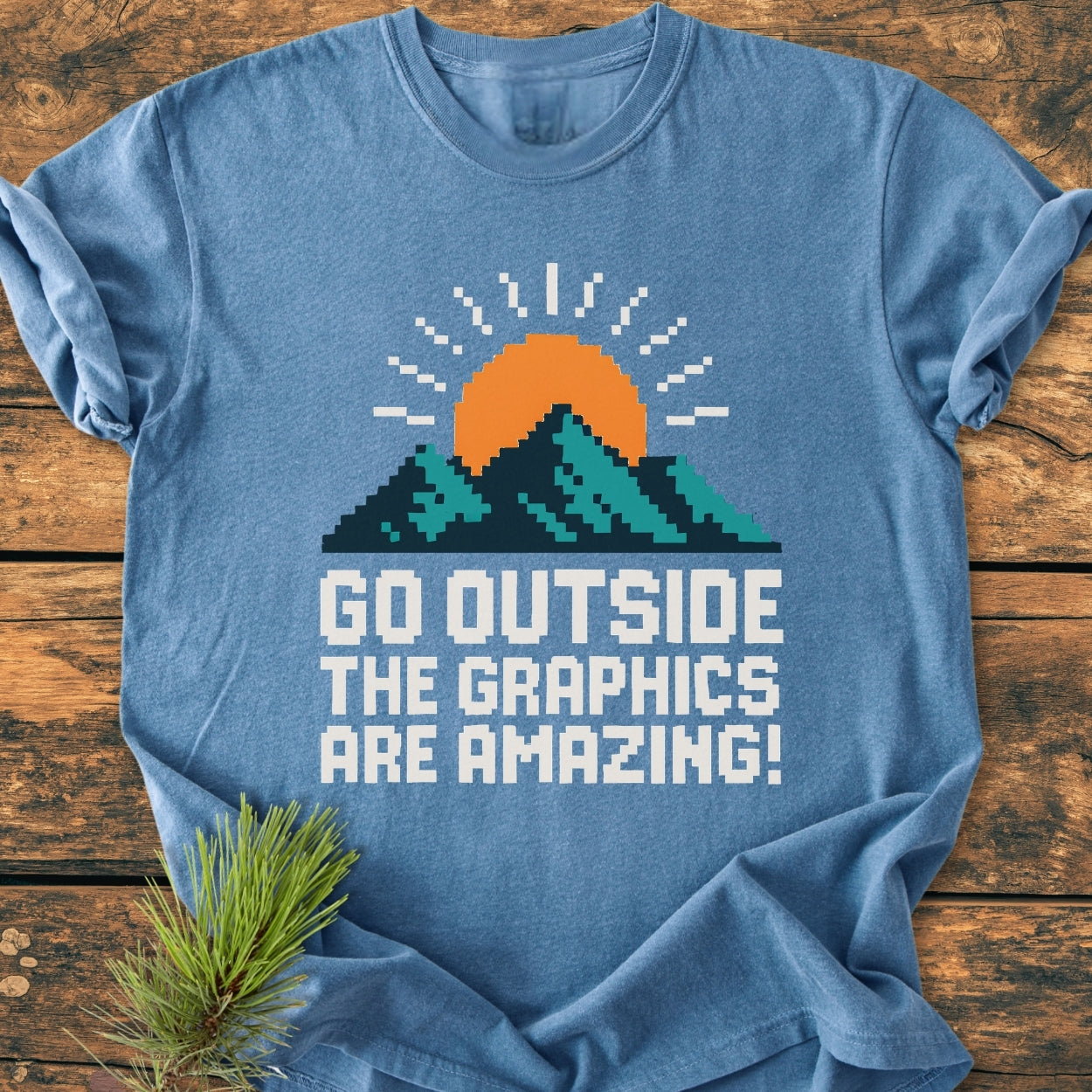 Heather-blue Go Outside Pixelart Mountain - Vintage Wash Tee featuring an 8-bit sun and mountain graphic, laid flat on wood with a pine sprig. Showcases chunky slogan, durable stitching, and relaxed fit.