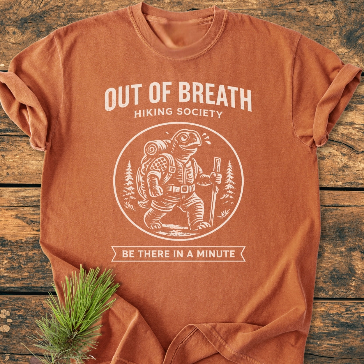 Out Of Breath Turtle - Vintage Wash Tee features a circular graphic of a turtle with a hiking staff, surrounded by trees, on a rust-orange background, complemented by the text, “OUT OF BREATH HIKING SOCIETY.”