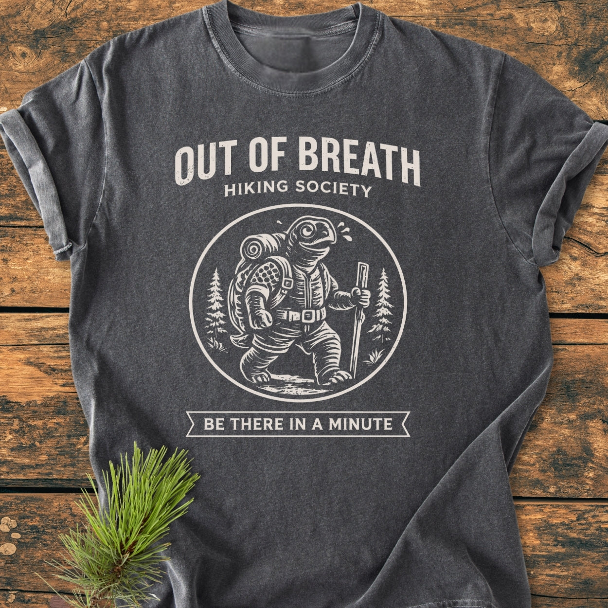 Out Of Breath Turtle - Vintage Wash Tee features a relaxed fit, charcoal-gray design with a circular badge of a hiking turtle, printed in off-white, laid on a wooden surface with a spruce sprig.