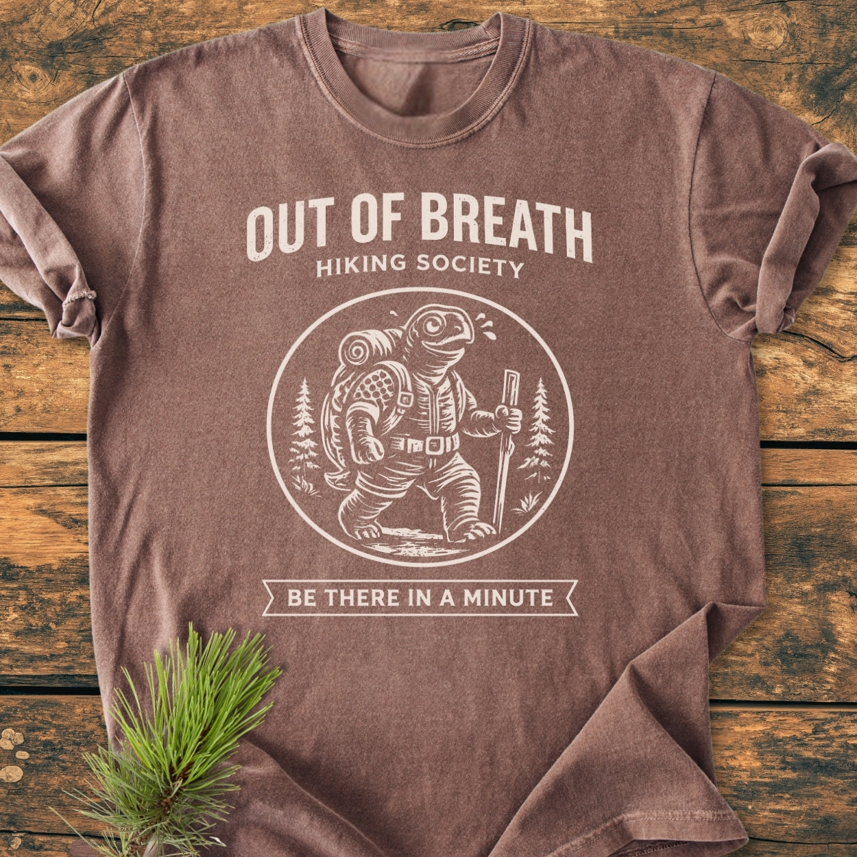 Out Of Breath Turtle - Vintage Wash Tee featuring an anthropomorphic turtle in hiking gear. The shirt is laid on a wooden surface with a pine sprig beside it, showcasing the hiking-themed graphic.