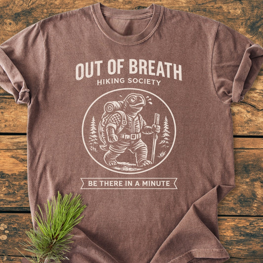 Out Of Breath Turtle - Vintage Wash Tee featuring an anthropomorphic turtle in hiking gear. The shirt is laid on a wooden surface with a pine sprig beside it, showcasing the hiking-themed graphic.