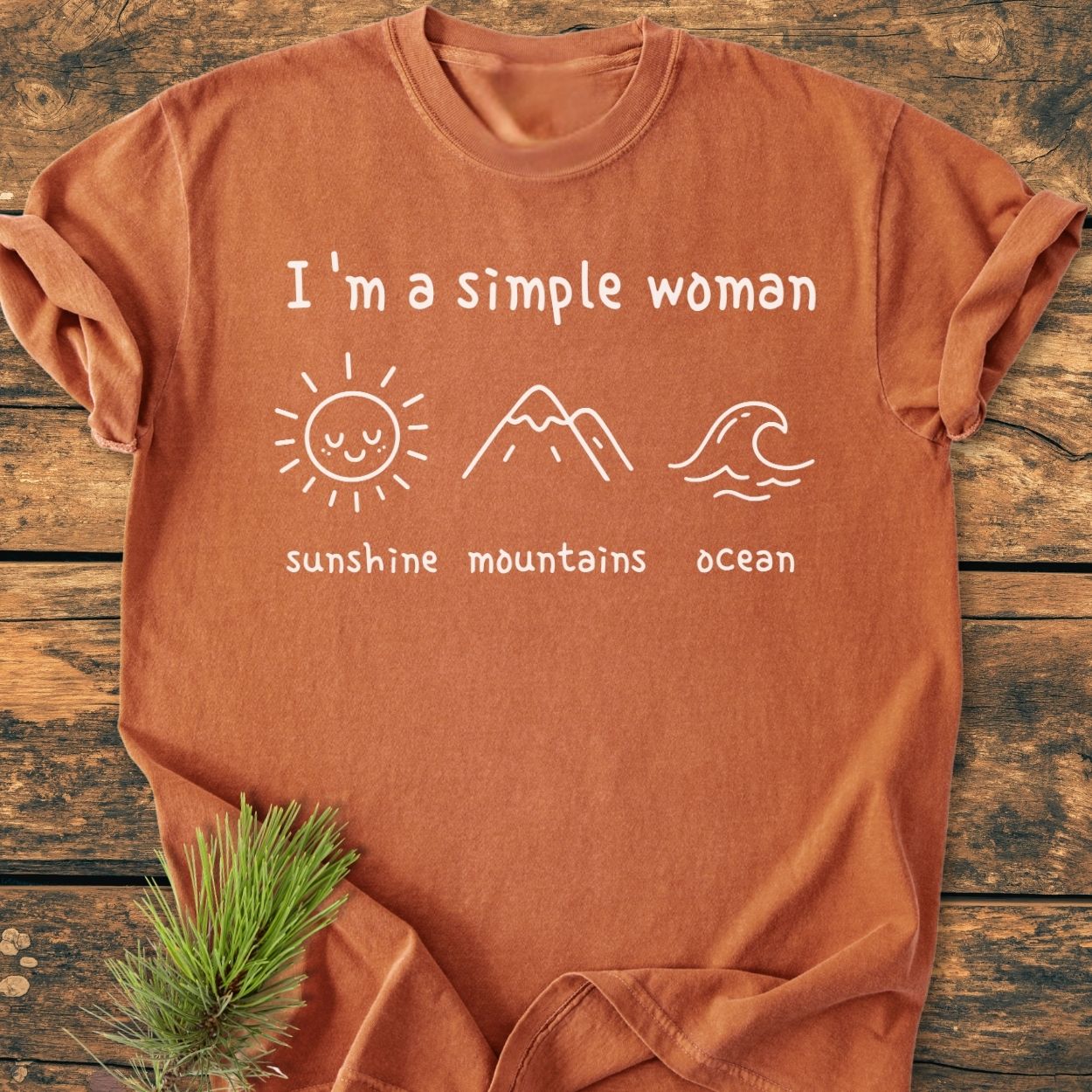 Burnt-orange tee with “I’m A Simple Woman. Sunshine Mountains Ocean” design, featuring line-drawn icons of a sun, mountains, and ocean. Laid on a wooden surface with a pine sprig beside it.