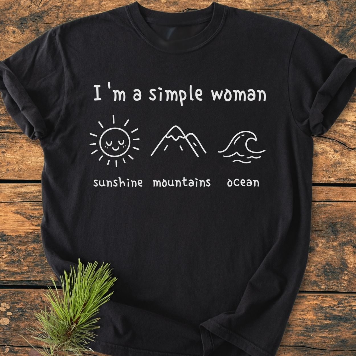 I'm A Simple Woman. Sunshine Mountains Ocean Vintage Wash Tee, black with white text and icons, laid flat on wood. Features a soft, relaxed fit, garment-dyed for comfort, and durable stitching.