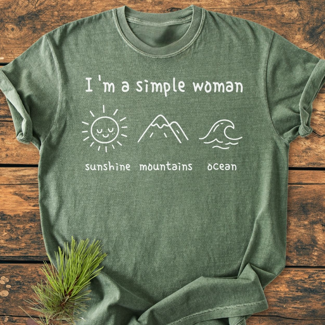I'm A Simple Woman. Sunshine Mountains Ocean - Vintage Wash Tee displayed flat on a wooden surface, featuring minimalist icons and text in white. The tee highlights a relaxed fit with durable, soft-washed fabric.