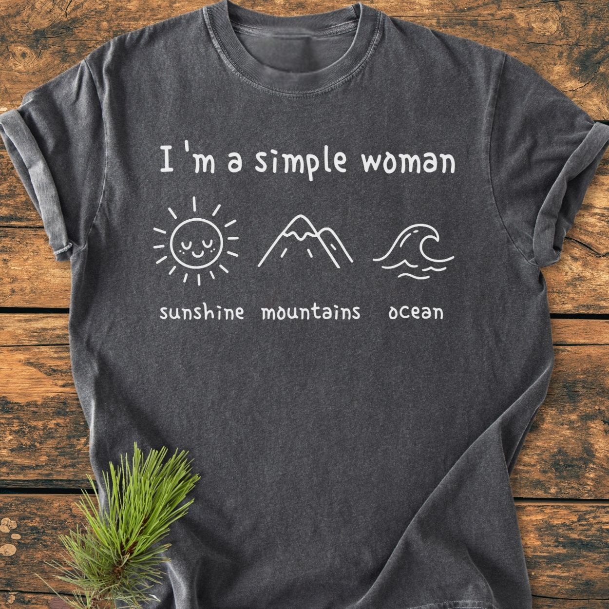 I'm A Simple Woman. Sunshine Mountains Ocean - Vintage Wash Tee features white lettering with sun, mountain, and wave icons on a charcoal-gray T-shirt, laid flat on a wooden surface.