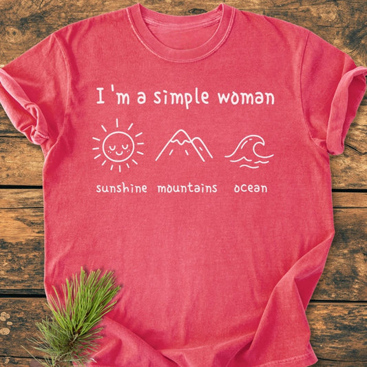 I'm A Simple Woman. Sunshine Mountains Ocean - Vintage Wash Tee featuring white lettering and icons of a sun, mountains, and ocean on a reddish-coral T-shirt, laid flat with a pine sprig accent.