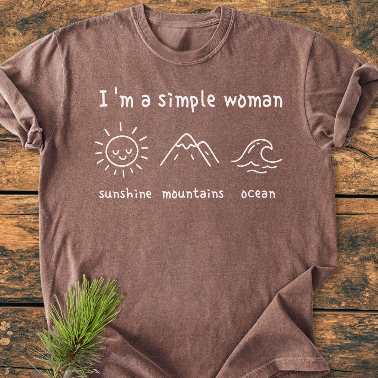 I'm A Simple Woman. Sunshine Mountains Ocean - Vintage Wash Tee: Brown crew-neck, soft-washed, featuring white handwritten text and line-drawings of a sun, mountains, and wave, laid flat on wood.