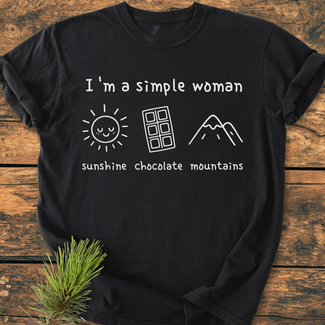 I'm A Simple Woman. Sunshine Chocolate Mountains - Vintage Wash Tee; black T-shirt with white print of sun, chocolate bar, mountain icons, laid on a wooden surface, emphasizing comfort and durability.