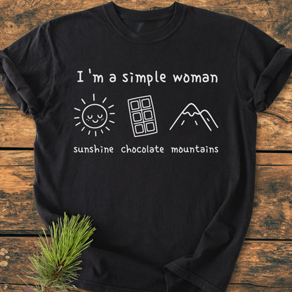 I'm A Simple Woman. Sunshine Chocolate Mountains - Vintage Wash Tee; black T-shirt with white print of sun, chocolate bar, mountain icons, laid on a wooden surface, emphasizing comfort and durability.