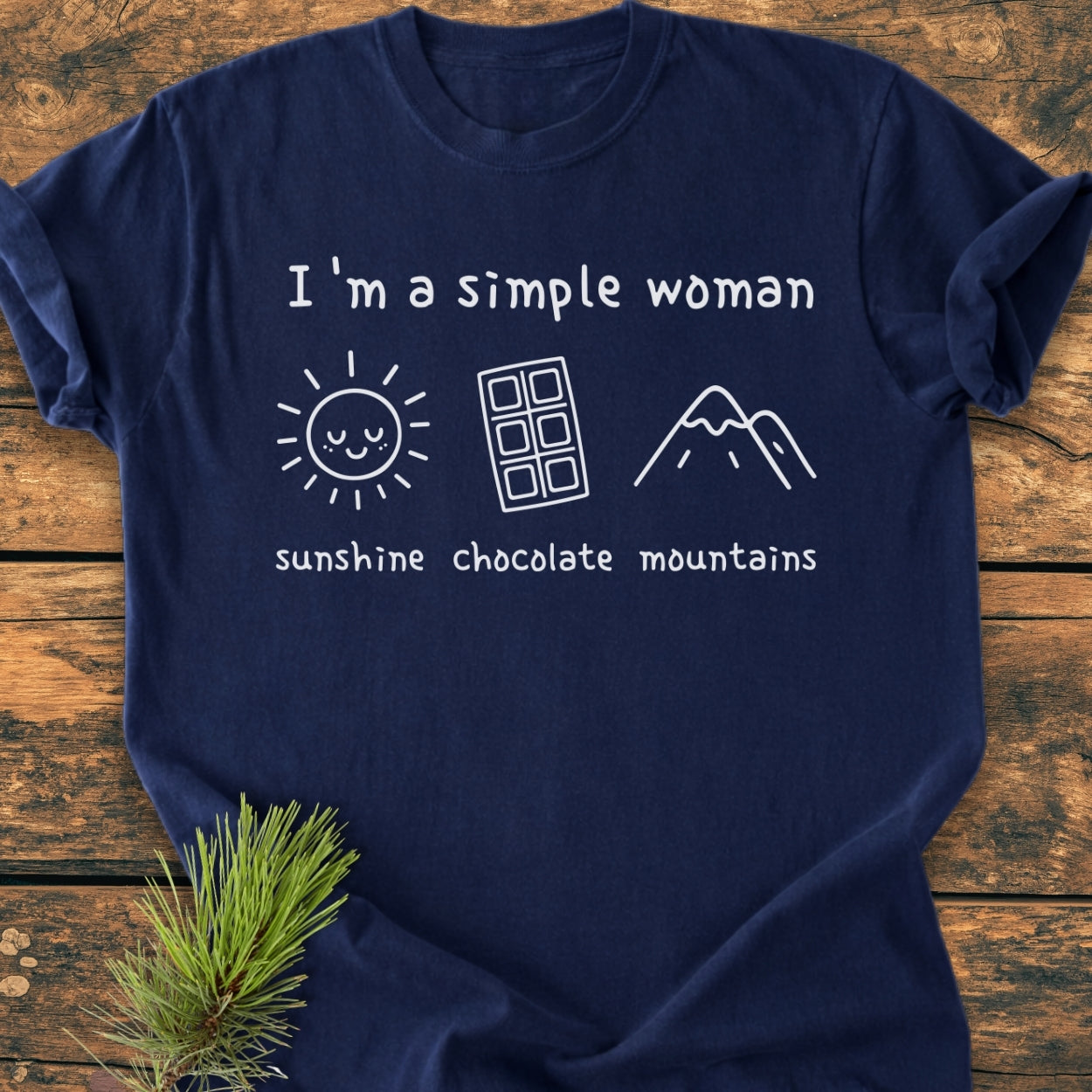 Navy-blue I'm A Simple Woman. Sunshine Chocolate Mountains - Vintage Wash Tee with white lettering and icons of a sun, chocolate bar, and mountains, laid flat on a wooden surface.