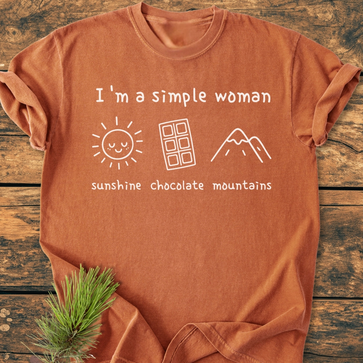 I'm A Simple Woman. Sunshine Chocolate Mountains - Vintage Wash Tee featuring a rust-orange finish with a slogan and icons of a sun, chocolate bar, and mountains, laid flat on wood.