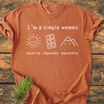 I'm A Simple Woman. Sunshine Chocolate Mountains - Vintage Wash Tee featuring a rust-orange finish with a slogan and icons of a sun, chocolate bar, and mountains, laid flat on wood.