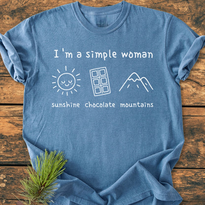 I'm A Simple Woman. Sunshine Chocolate Mountains - Vintage Wash Tee in blue, laid flat with white icons of a sun, chocolate bar, and mountains. Garment-dyed, soft-washed cotton, relaxed fit. Pine sprig accent.