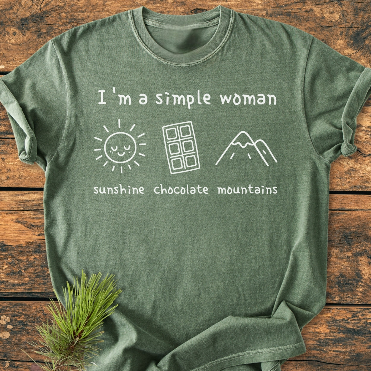 I'm A Simple Woman. Sunshine Chocolate Mountains - Vintage Wash Tee featuring white print on green, with icons of a sun, chocolate bar, and mountain, laid on a wooden surface.