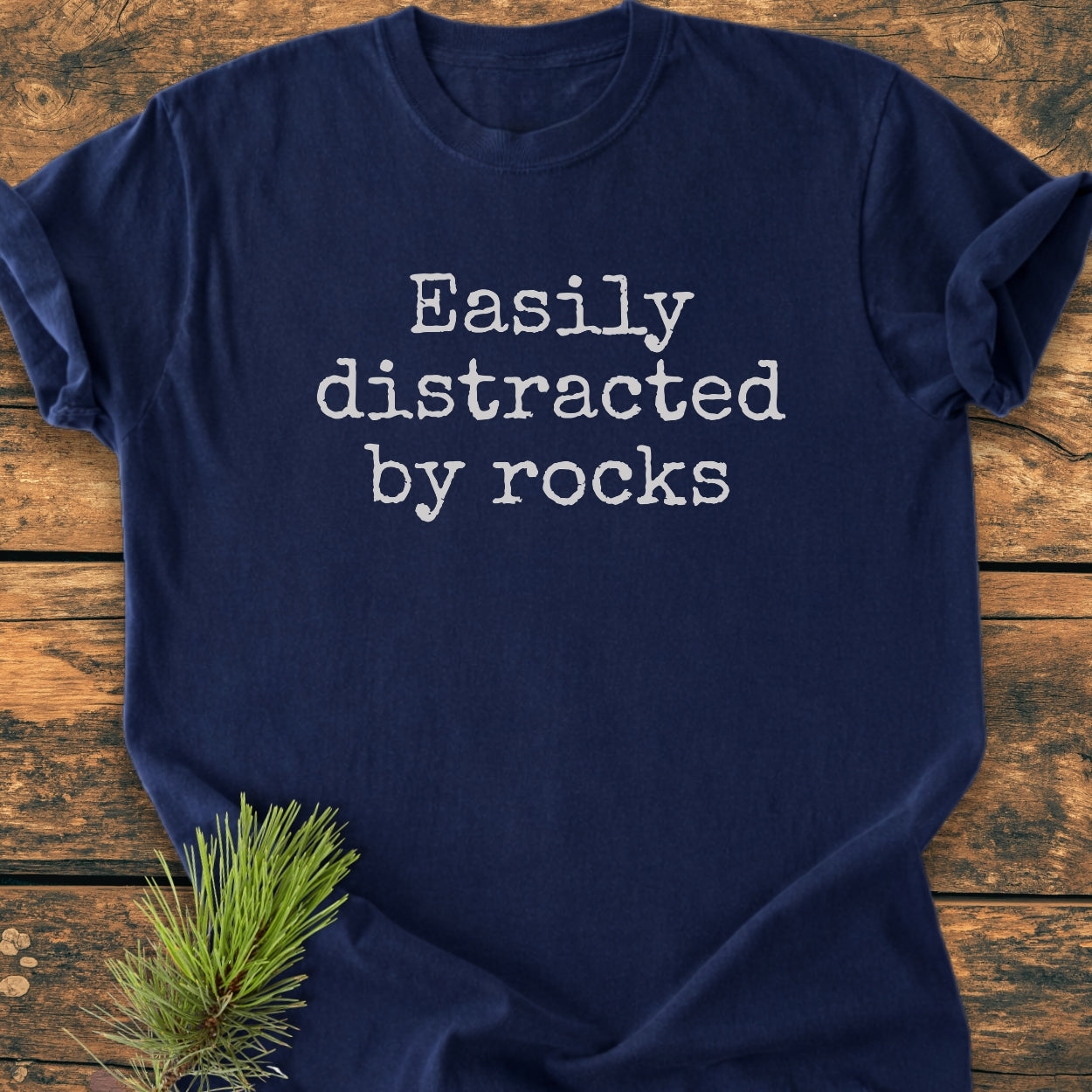 Navy-blue Easily Distracted By Rocks - Vintage Wash Tee with off-white typewriter font, laid flat on rustic wood, featuring a pine needle sprig. Cozy, relaxed fit, durable with double-needle stitching.