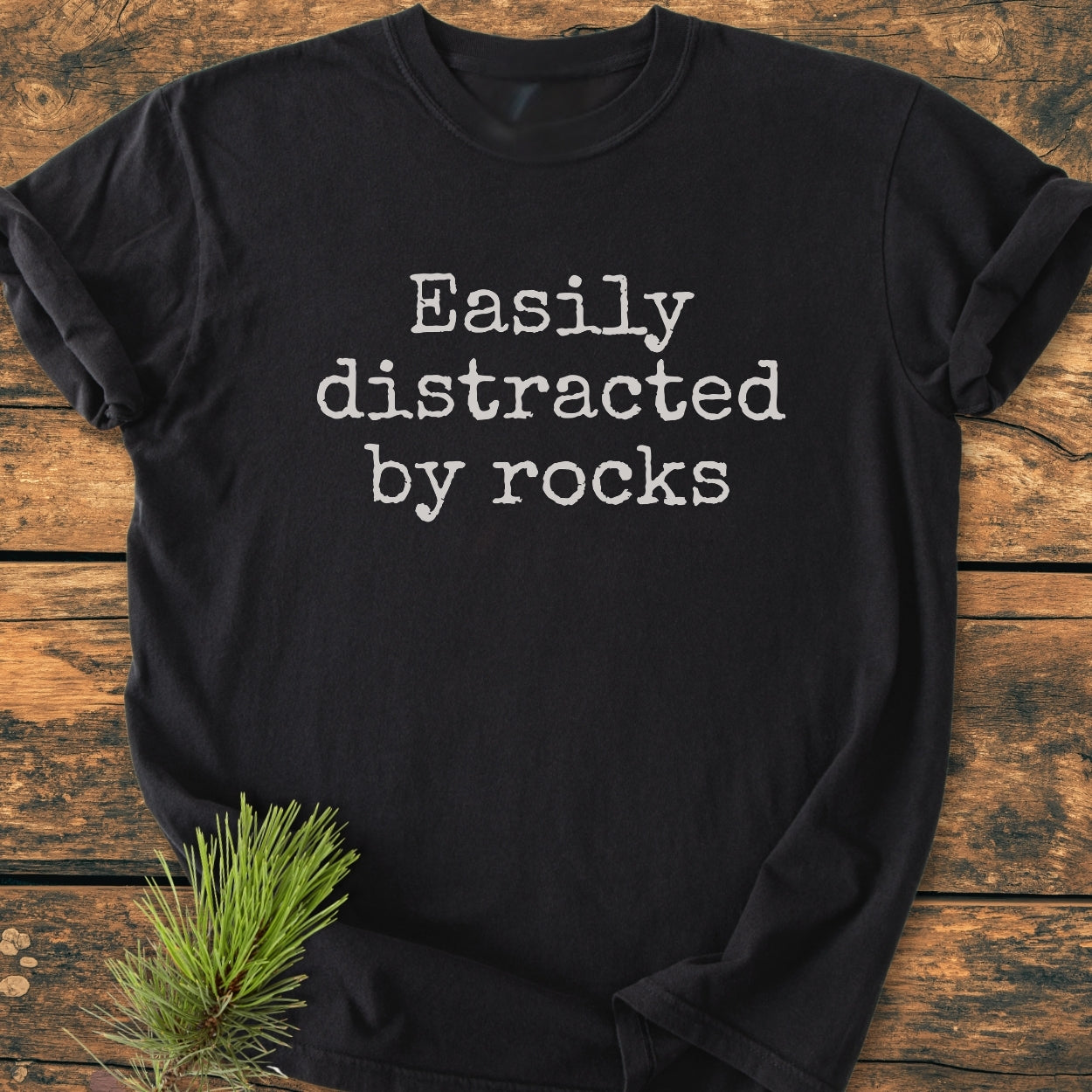 Black Easily Distracted By Rocks - Vintage Wash Tee on wooden surface, typewriter-style text, rolled sleeves, pine sprig detail. Crafted from soft, durable, pre-shrunk cotton with a relaxed fit.