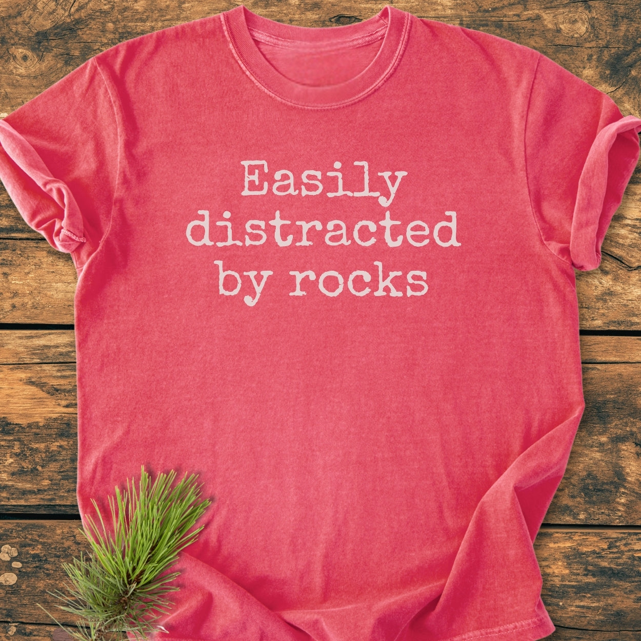 Easily Distracted By Rocks - Vintage Wash Tee displayed on wood, features white text and a pine sprig accent. Made from 100% ring-spun cotton, offering a relaxed fit and durable, soft-washed fabric.