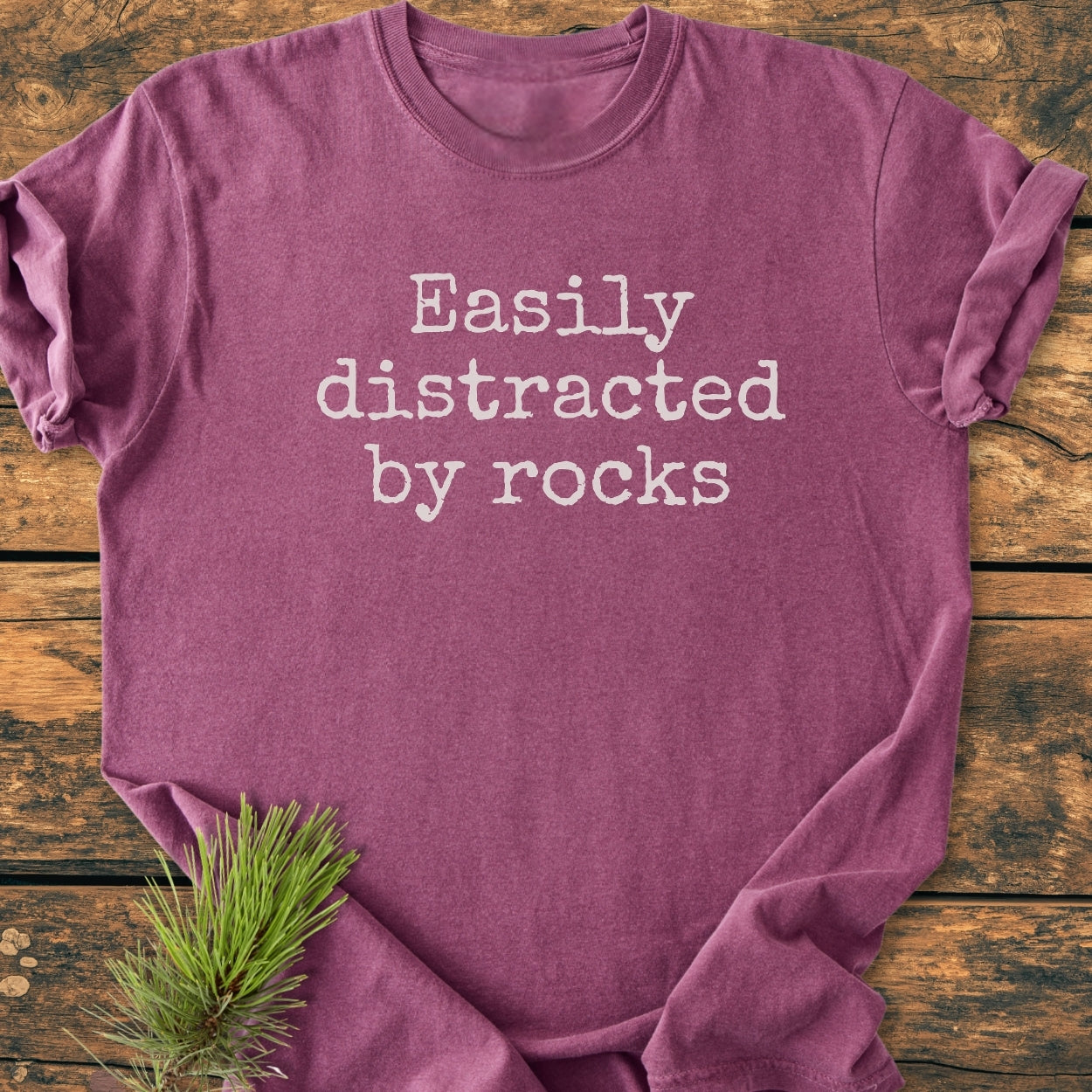 Easily Distracted By Rocks - Vintage Wash Tee, heather-purple, features typewriter-style text. Laid on wood with a pine sprig, showcasing Comfort Colors' soft, durable, ring-spun cotton design.