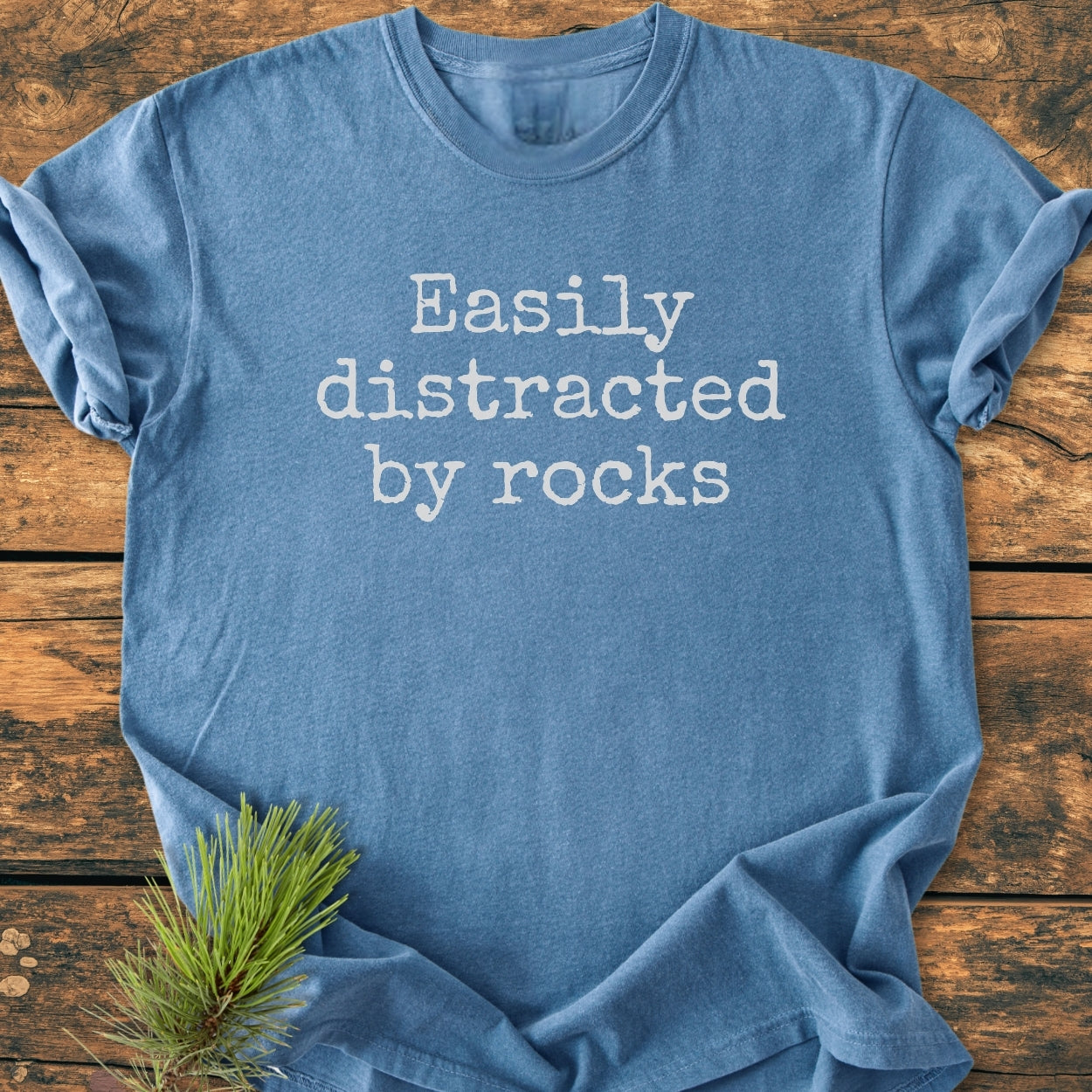 Easily Distracted By Rocks - Vintage Wash Tee on rustic wood, featuring white typewriter-style text. Laid flat, highlighting its relaxed fit and soft, garment-dyed fabric with a touch of pine needles.