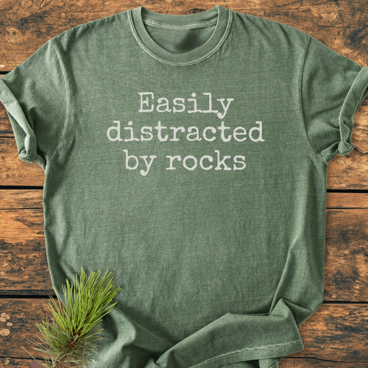 Easily Distracted By Rocks - Vintage Wash Tee in heather-forest green, features a typewriter-style font. Laid on wood, with a pine sprig accent. Cozy, durable, relaxed fit, 100% ring-spun cotton.