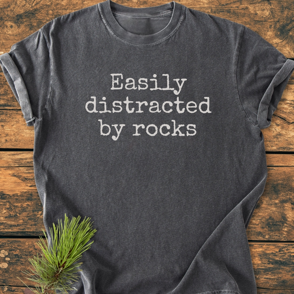Easily Distracted By Rocks - Vintage Wash Tee: Dark gray T-shirt with off-white typewriter font, laid flat with rolled sleeves on wood. Pine needles add a rustic touch, enhancing its outdoorsy appeal.