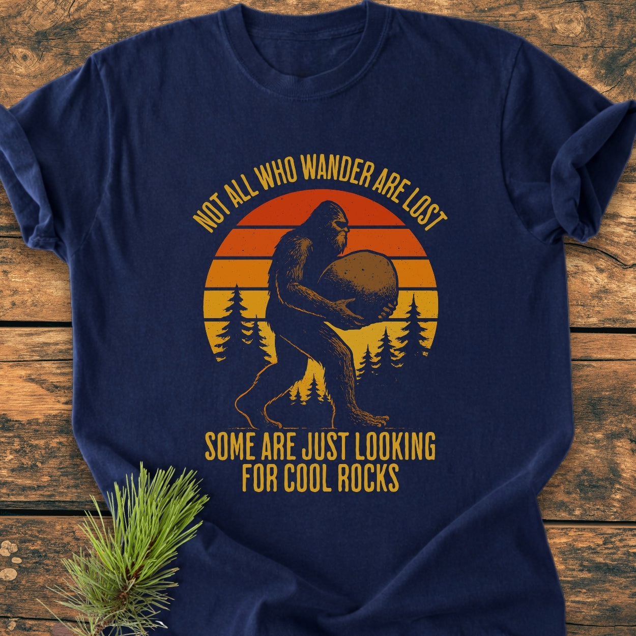 Looking For Cool Rocks - Vintage Wash Tee features Bigfoot carrying a stone with pine trees and a sunset backdrop. This comfortable, navy-blue tee offers a relaxed fit and durable double-needle stitching.