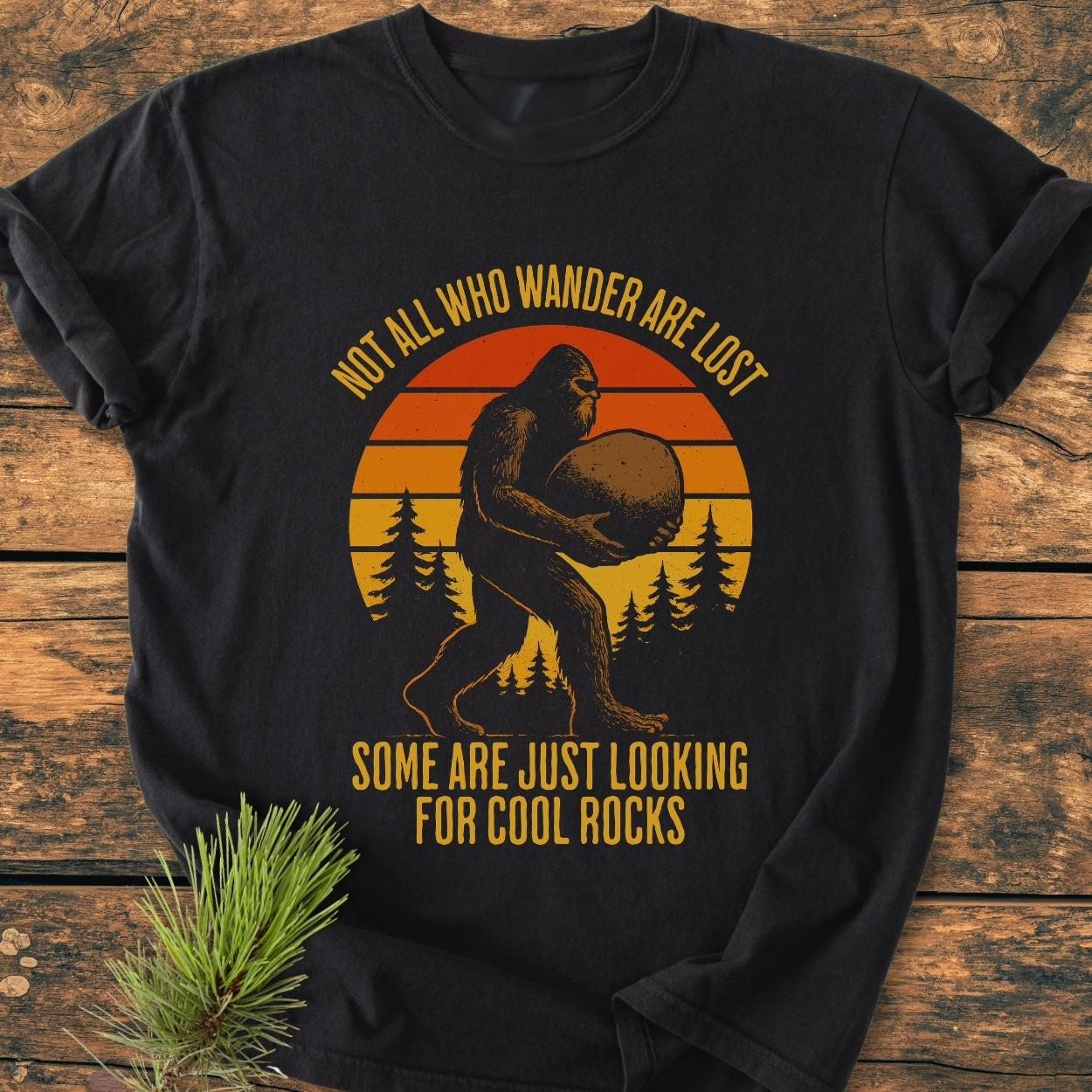 Looking For Cool Rocks - Vintage Wash Tee: Black T-shirt with a vintage Bigfoot graphic carrying a rock, set against a sunset and pine trees, featuring a humorous slogan about wandering.