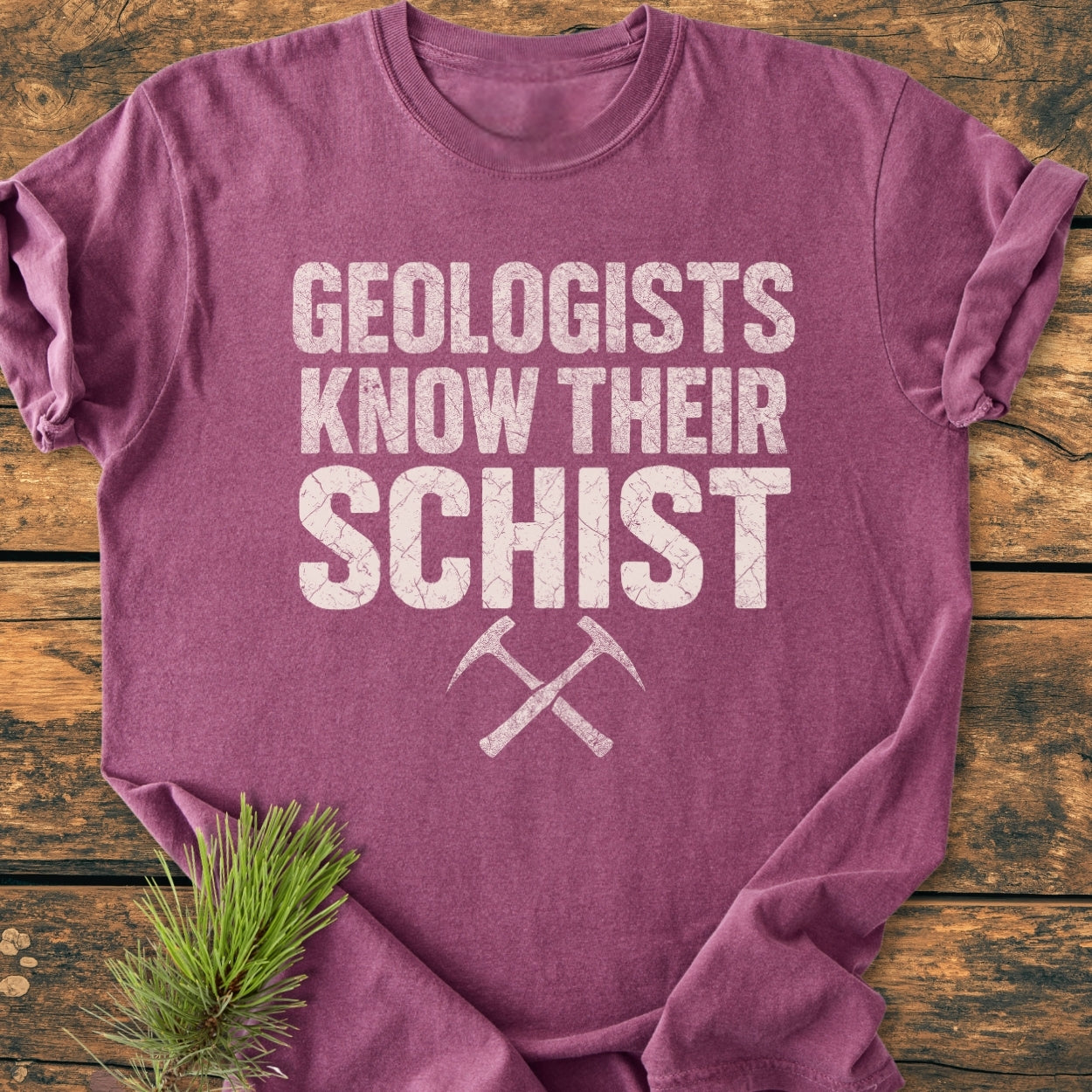 Geologists Know Their Schist - Vintage Wash Tee features white lettering and crossed rock-hammers on a mauve tee, laid flat on wood, with a pine sprig nearby.