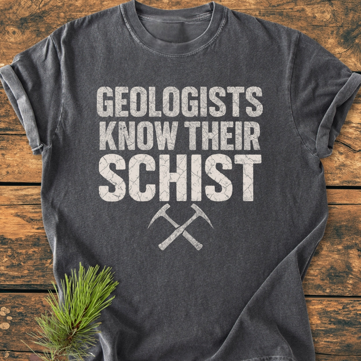 Geologists Know Their Schist - Vintage Wash Tee: Dark-gray, 100% ring-spun cotton, featuring Geologists Know Their Schist text and crossed rock-hammers, laid on wood with a pine sprig accent.