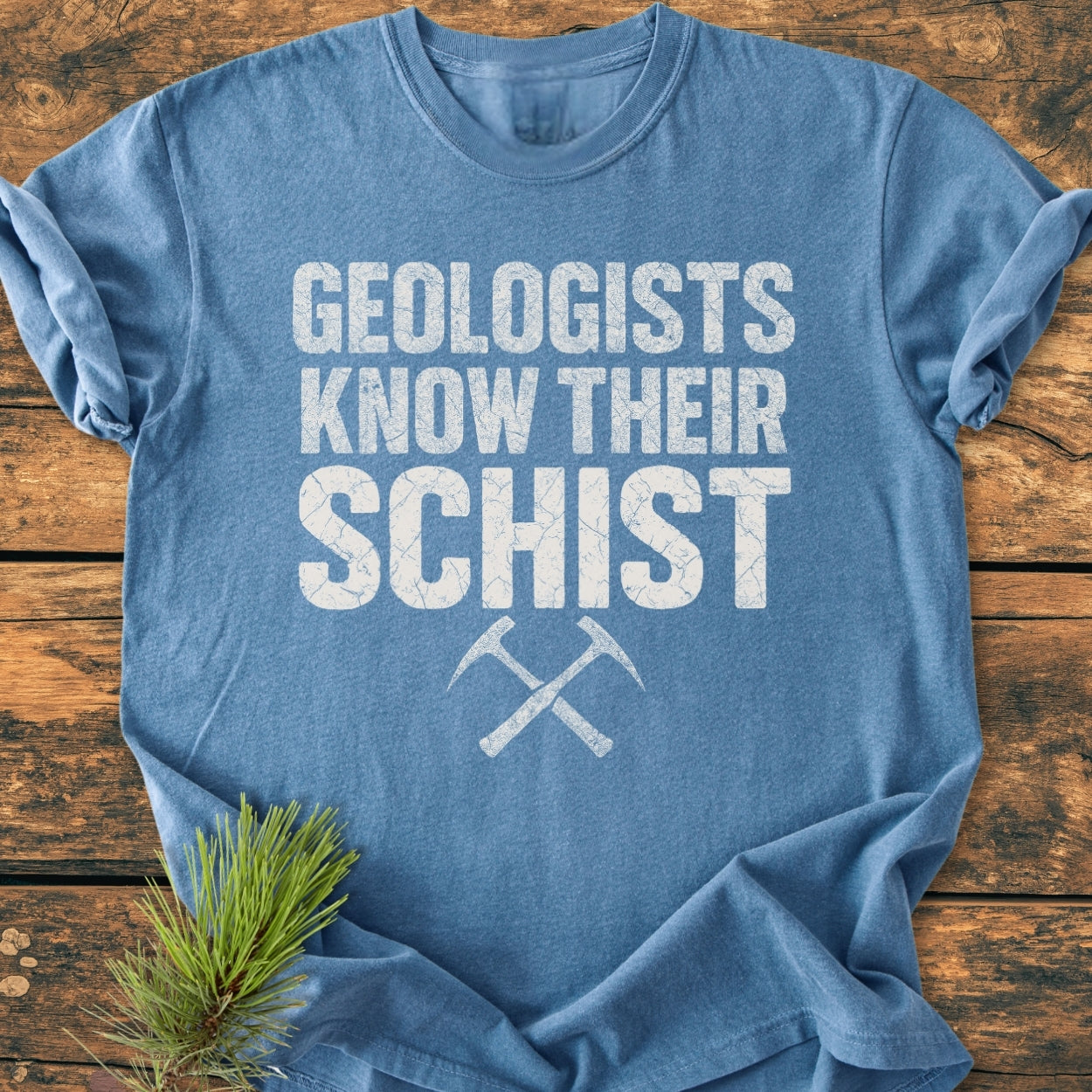 Geologists Know Their Schist - Vintage Wash Tee: Blue t-shirt with slogan in distressed white, crossed rock-hammer icons, on wooden surface. Pine sprig accentuates the soft, relaxed, durable garment.