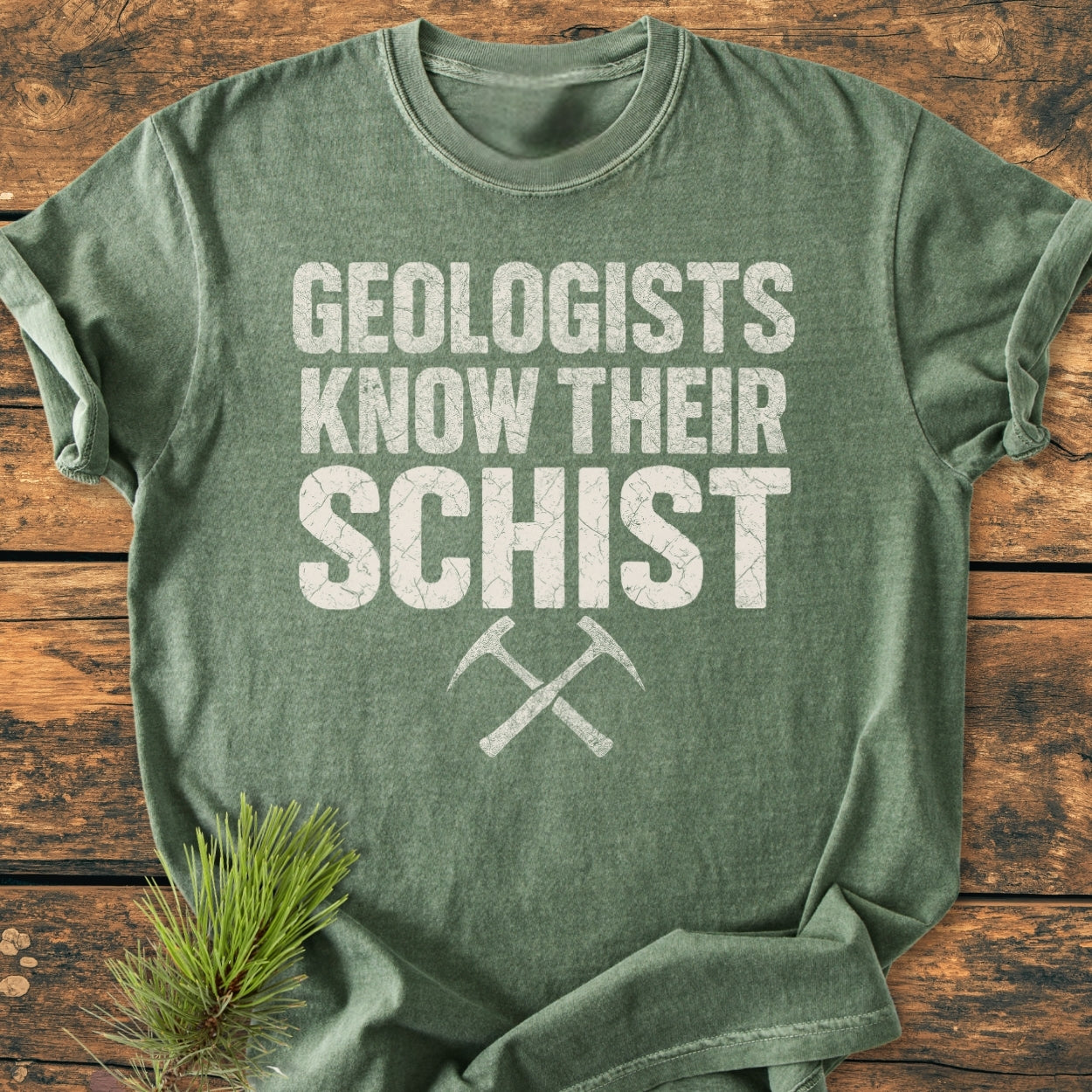 Geologists Know Their Schist - Vintage Wash Tee in olive-green with white weathered slogan and crossed rock-hammers graphic, displayed on wood with pine sprig; soft, relaxed, durable garment-dyed cotton.