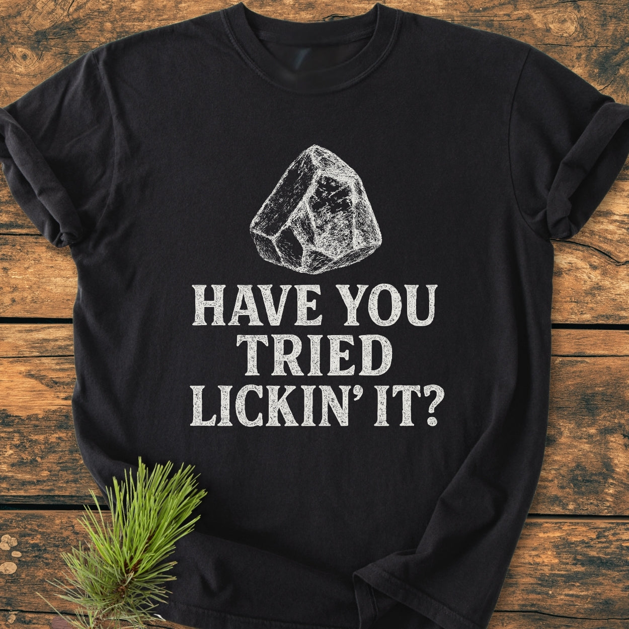Black t-shirt titled Have You Tried... Rockhound - Vintage Wash Tee featuring a white line-drawing of an ice cube and the phrase HAVE YOU TRIED LICKIN’ IT? styled with a pine branch accent.