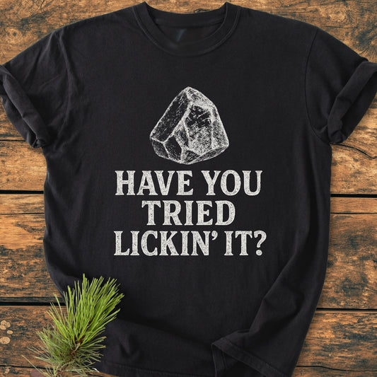 Black t-shirt titled Have You Tried... Rockhound - Vintage Wash Tee featuring a white line-drawing of an ice cube and the phrase HAVE YOU TRIED LICKIN’ IT? styled with a pine branch accent.