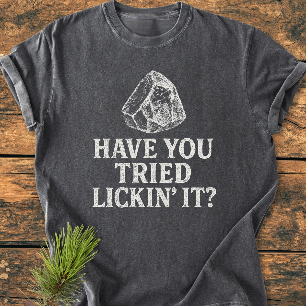 Have You Tried Lickin' It? Rockhound - Vintage Wash Tee featuring a white sketch of a rock and playful text on a dark-gray background, laid flat on a wood-grain surface with a pine sprig nearby.