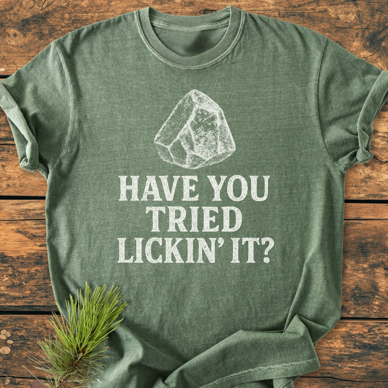 Forest-green Have You Tried... Rockhound - Vintage Wash Tee with a chalk-style sketch of a rock and the phrase, Have you tried lickin’ it? displayed on a wooden surface beside a pine sprig.