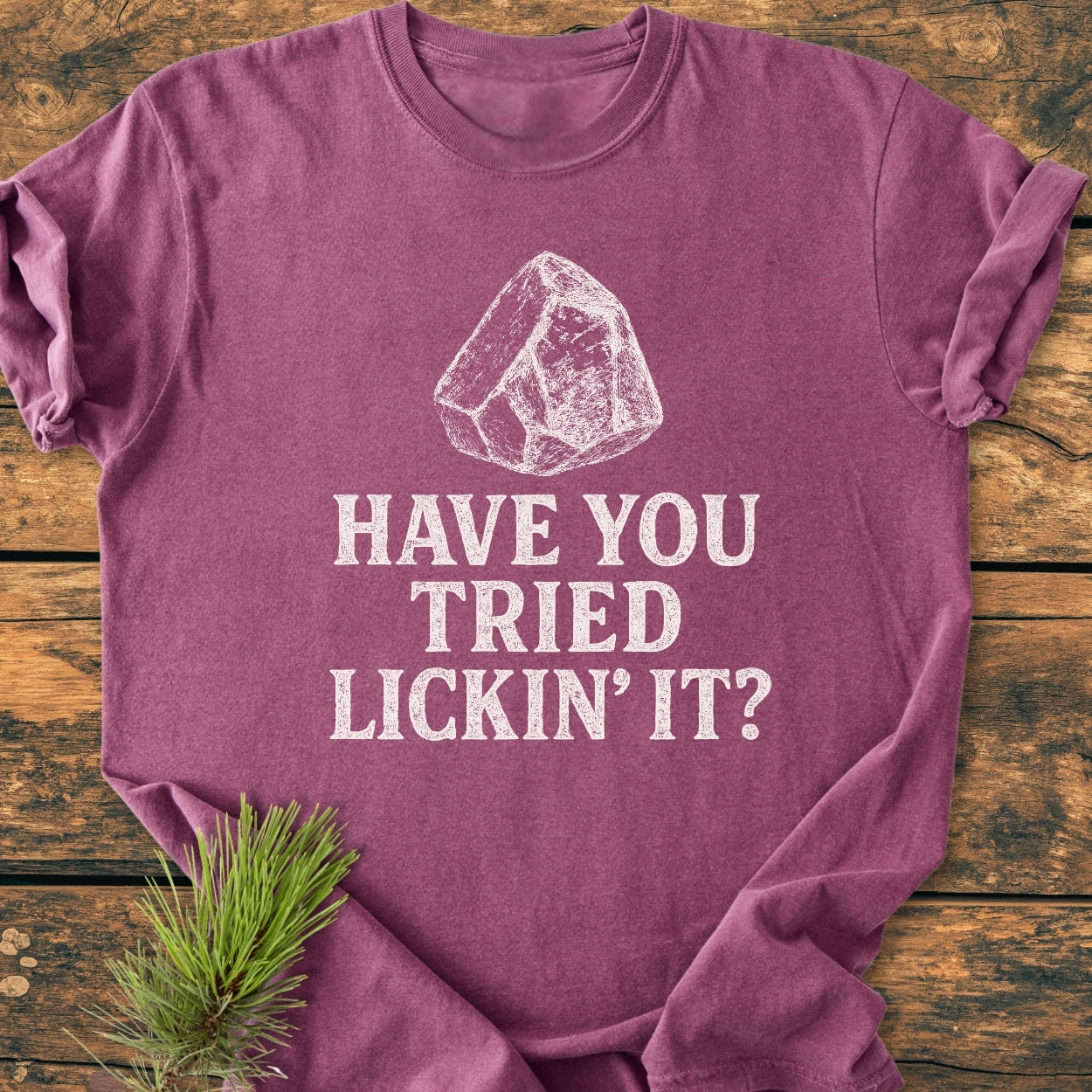 The Have You Tried Lickin' It? Rockhound - Vintage Wash Tee features a white line-drawing of a rock with a slogan, displayed on a heather-purple T-shirt with a pine sprig accent.