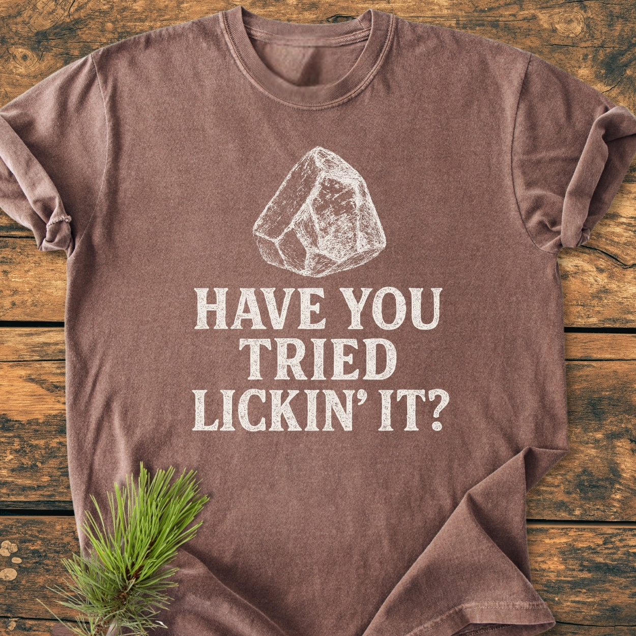 Brown Have You Tried Lickin' It? Rockhound - Vintage Wash Tee with white, chalk-style ice chunk sketch, laid on wood surface with pine sprig accent, showcasing its relaxed fit and soft-washed texture.