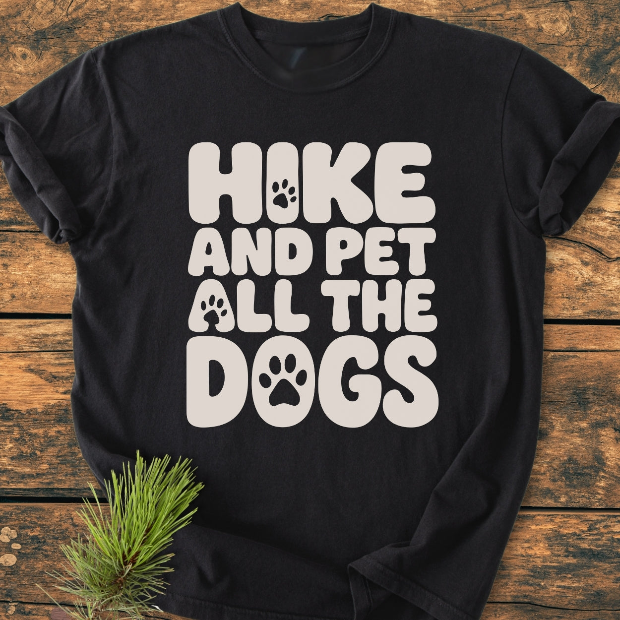 Hike And Pet All The Dogs - Vintage Wash Tee features cream bubble letters with paw-print motifs on a black T-shirt, laid flat on wood, accented by a small pine twig.