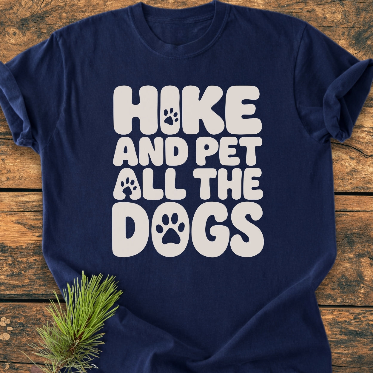 Navy-blue Hike And Pet All The Dogs - Vintage Wash Tee on wood. Features white slogan with paw-print graphics. Accompanied by a pine sprig, highlighting the shirt's cozy, relaxed fit.
