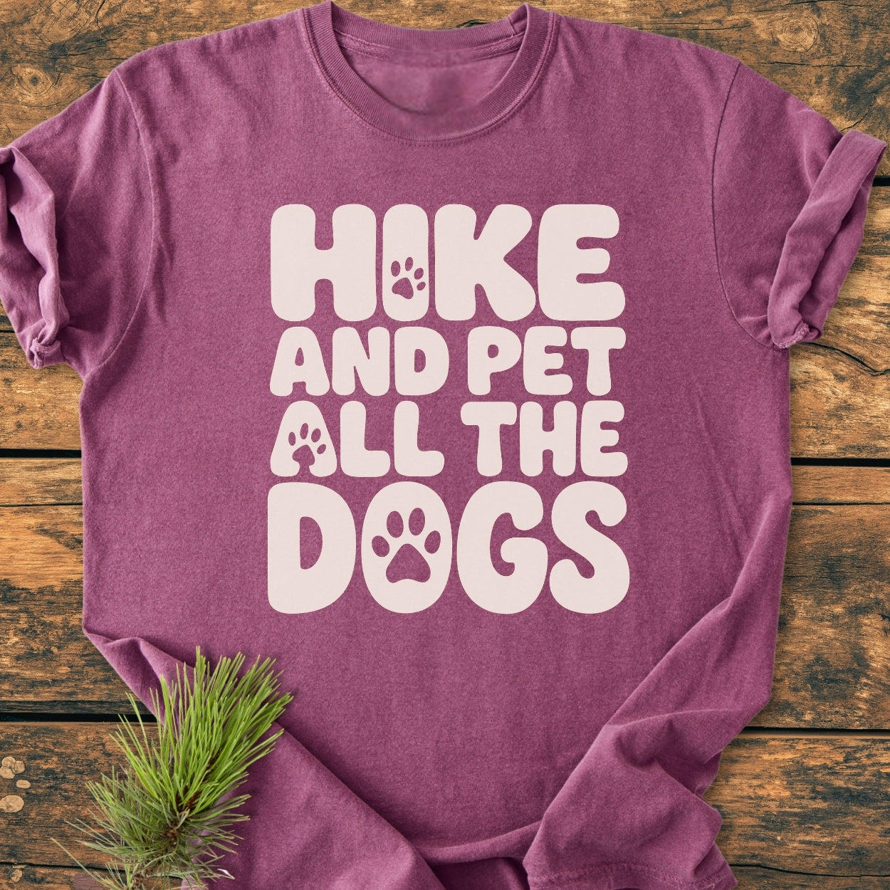 Hike And Pet All The Dogs - Vintage Wash Tee in plum color, featuring cream slogan with paw-print details, laid on wood. Includes pine sprig accent, emphasizing cozy, relaxed fit and durability.