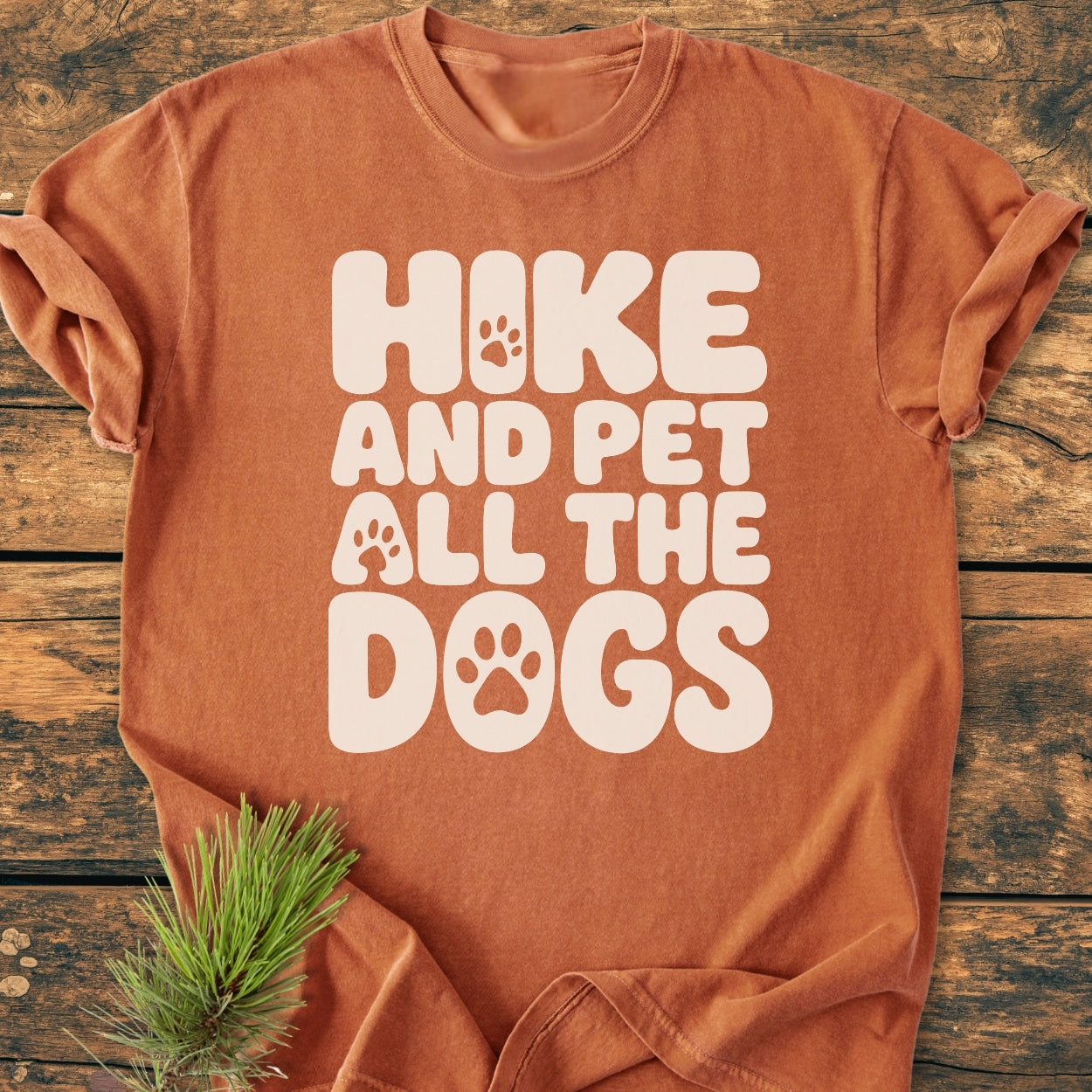 Rust-orange Hike And Pet All The Dogs - Vintage Wash Tee with cream bubble letters, paw-print details on a wooden surface beside a pine sprig, showcasing comfort and durability.