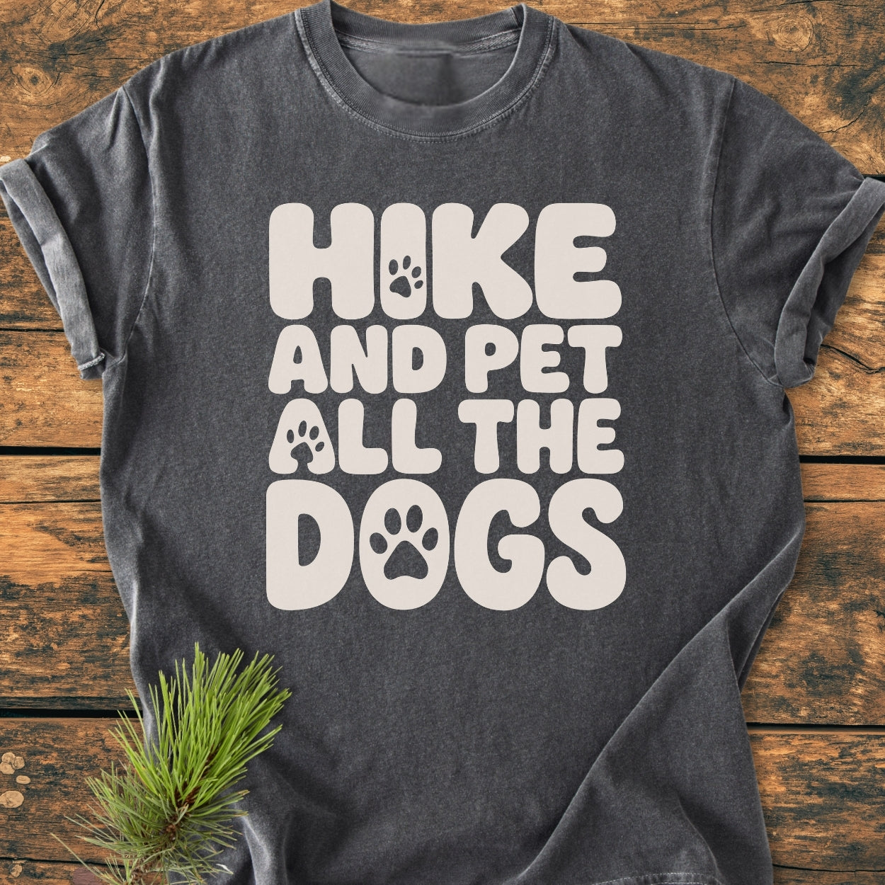 Hike And Pet All The Dogs - Vintage Wash Tee featuring a chunky cream slogan with paw prints on a charcoal-gray background, laid flat on wood, with a pine sprig accentuating its cozy, relaxed fit.