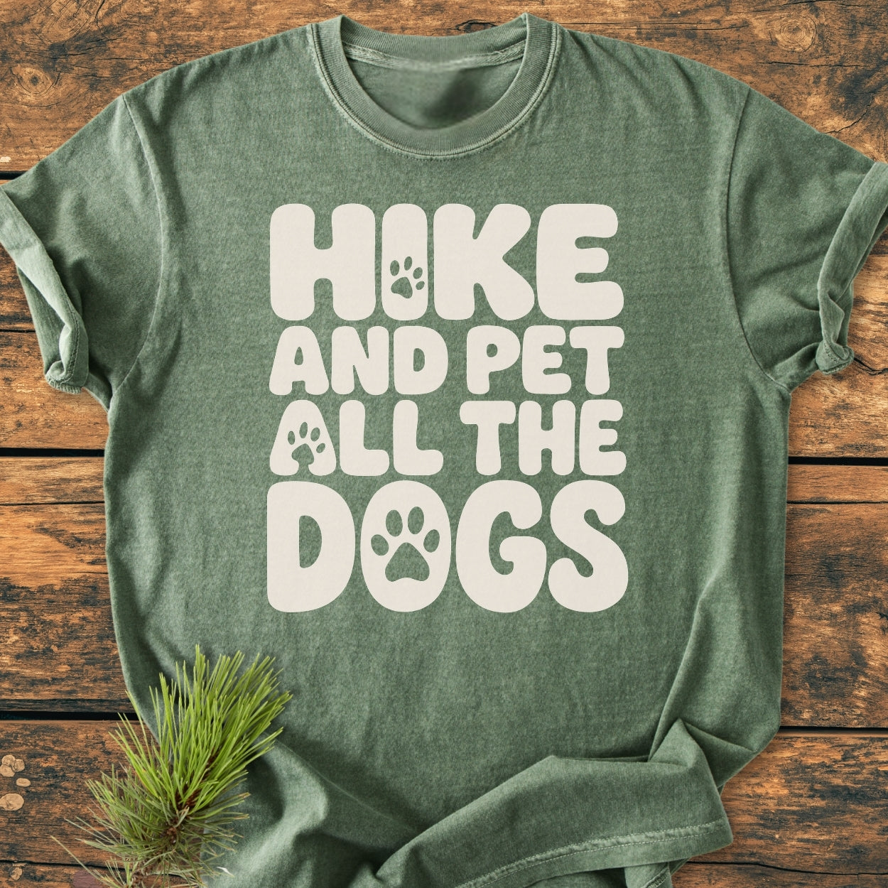 Heather-green Hike And Pet All The Dogs - Vintage Wash Tee with playful paw-print design and white text, laid flat on wood with a pine branch, showcasing its relaxed fit and soft texture.